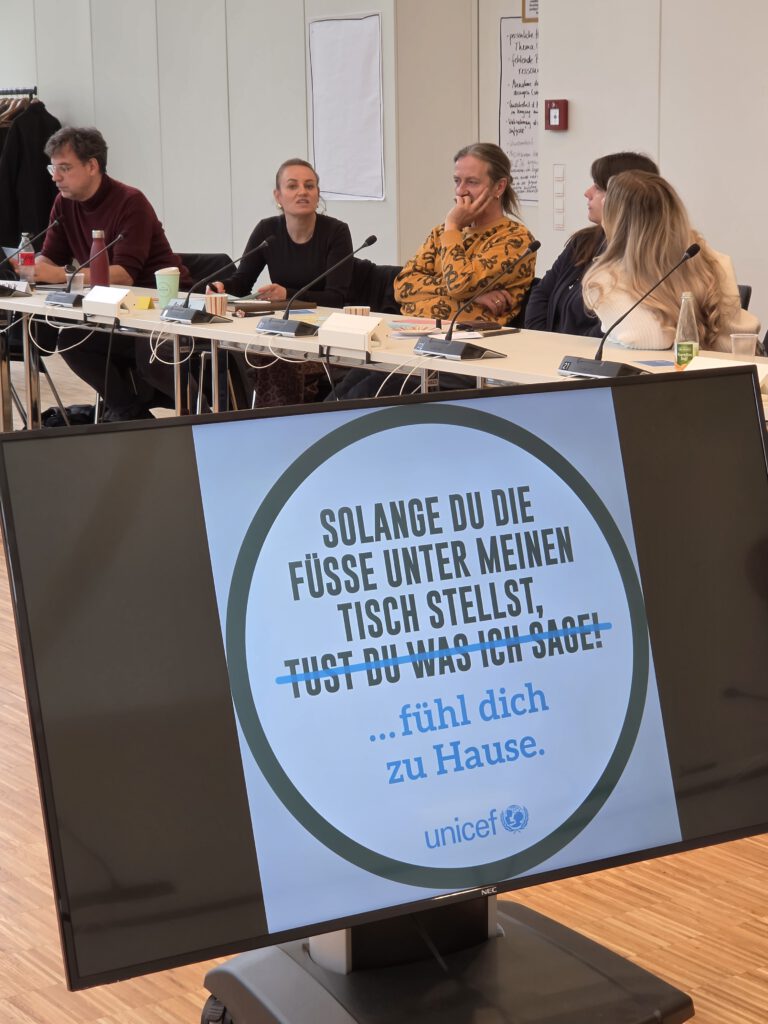 Personen sitzen an Tischen hinter einem Monitor mit einem Slogan von unicef: Solange du die Füße unter meinen Tisch stelllst, fühl dich zuhause.