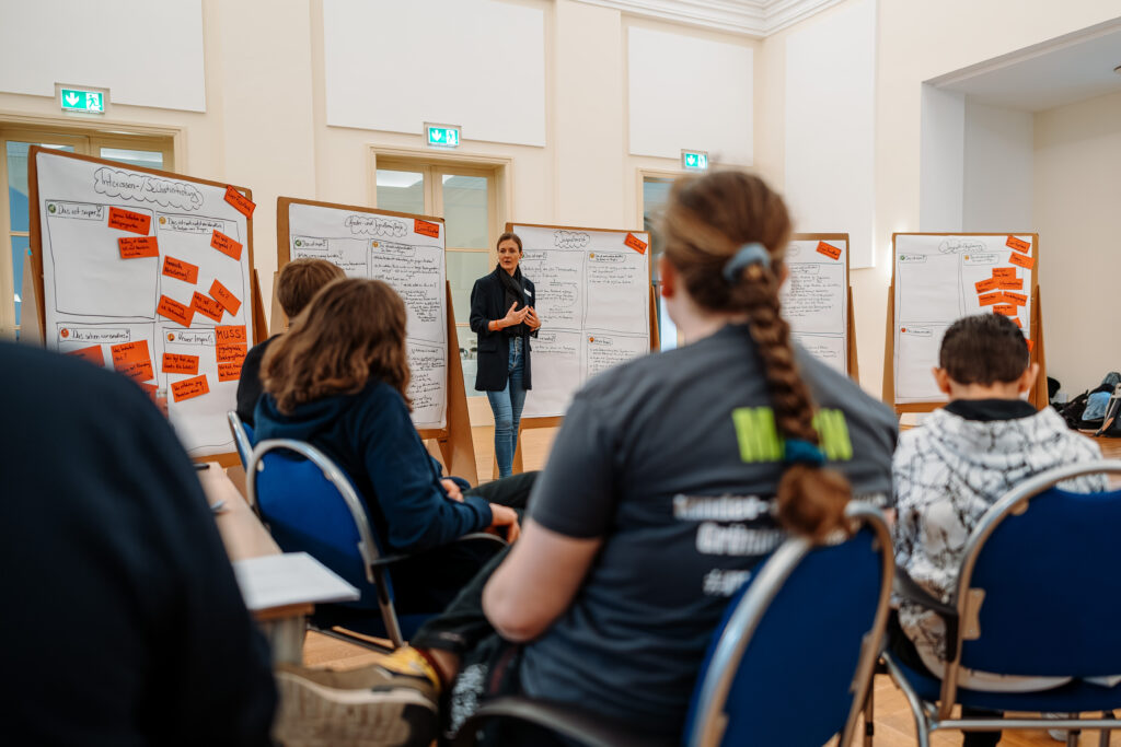 Mehrere Personen sitzen in einem Workshopraum; vorne steht eine Person zwischen Stellwänden mit roten Moderationskarten.