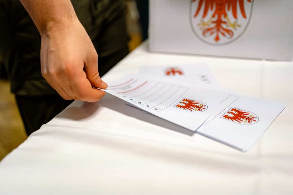 Eine Hand hält einen Papierbogen über Unterlagen mit dem roten Brandenburger Adler.